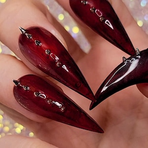 Puede incluir: Primer plano de uñas artificiales largas y puntiagudas, de color burdeos. Cada uña está adornada con pequeños tachuelas plateadas y una punta plateada en la punta. Las uñas tienen un acabado brillante y están dispuestas en forma de abanico.