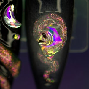 Puede incluir: Dos puntas de uñas negras con purpurina iridiscente y un diseño de media luna. La purpurina es una mezcla de oro, rosa y morado.