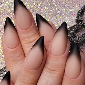 Puede incluir: Primer plano de una mano que muestra uñas largas en forma de estilete con un diseño degradado de negro a nude. Las uñas tienen un acabado mate, con el color negro desvaneciéndose en un nude natural en la base. El fondo es una superficie brillante e iridiscente.