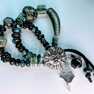 May include: A multi-strand bracelet with green, brown, and silver beads. The bracelet features a silver sun charm with a face and a silver bird charm with a crystal bead.