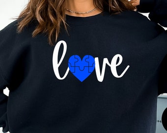 May include: Black sweatshirt with the word "love" in white script, featuring a blue puzzle-piece heart. The word "BLACK" is in a white circle in the lower right corner. The sweatshirt is a casual style.