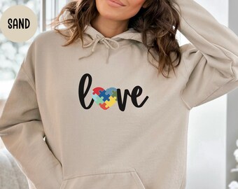 May include: Sand-colored hoodie with a front pocket and drawstring. The word "love" is written in black script, with the "o" replaced by a heart made of colorful puzzle pieces. The word "SAND" is in the top left corner.
