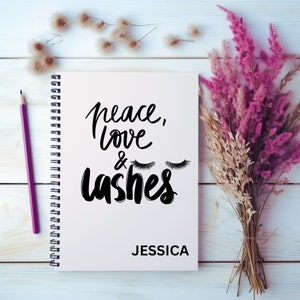 May include: A white spiral-bound notebook with the words "peace, love & lashes" in black script, along with a pair of eyelash illustrations. The name "JESSICA" is printed below. A purple pencil and a bouquet of pink and beige dried flowers are also visible.