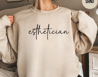Op de afbeelding: Zandkleurige sweatshirt met het woord "esthetician" in zwart script. De top met lange mouwen is gemaakt van een zacht materiaal. De sweatshirt heeft een neutrale kleur en is geschikt voor vrijetijdskleding.