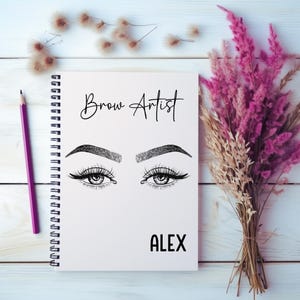 Op de afbeelding: Een witte spiraalgebonden notitieboek met de woorden "Brow Artist" en de naam "ALEX" op de kaft. De kaft heeft een gedetailleerde illustratie van een paar ogen met lange wimpers en perfect gevormde wenkbrauwen. Een paars potlood en droogbloemen staan ook in de afbeelding.