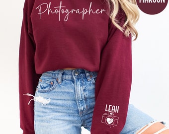 May include: Maroon sweatshirt with the word "Photographer" in white script across the chest. The right sleeve features the name "LEAH" and a camera graphic with a heart. The sweatshirt is a solid maroon color.