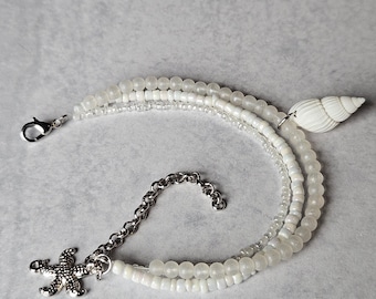 May include: A white beaded bracelet with a silver starfish charm and a white seashell charm. The bracelet is made of three strands of beads.
