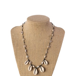 May include: A necklace made of cowrie shells and black cord. The necklace is displayed on a brown fabric mannequin.