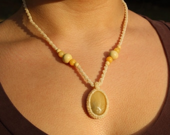 May include: Cream-colored macrame necklace with a pendant and wooden beads. The oval pendant is a light yellow color, and the necklace is worn against a brown top. The necklace features a woven design.