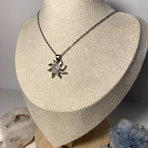 May include: A silver chain necklace with a sun pendant. The pendant is a stylized sun with a circular center and radiating rays.