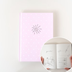 May include: A pink journal with white polka dots and a black line drawing of a burst on the cover. The open pages show a line drawing of a desk and pens, with the word "goals" written in cursive.