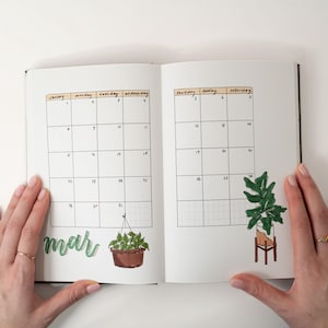 May include: Open notebook with a March calendar layout. The calendar grid is filled with dates and has illustrations of potted plants. The word "mar" is written in green cursive. The notebook is open, held by hands with gold rings.