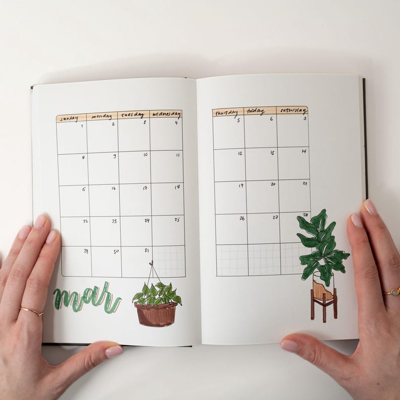 May include: Open notebook with a March calendar layout. The calendar grid is filled with dates and has illustrations of potted plants. The word "mar" is written in green cursive. The notebook is open, held by hands with gold rings.