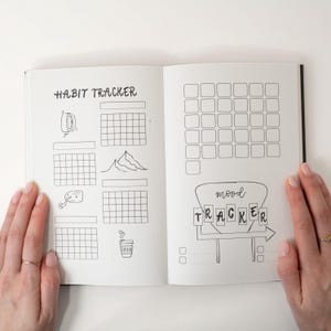 May include: Open journal with black cover, featuring habit and mood tracker pages. The left page has "HABIT TRACKER" with boxes and illustrations. The right page has a grid for tracking and "mood TRACKER" with an arrow.