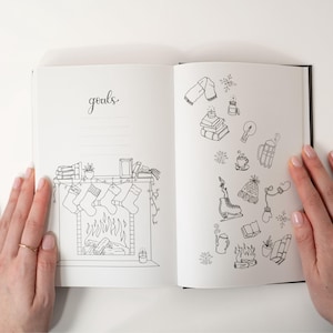 May include: Open journal with black cover, featuring hand-drawn illustrations. One page has the word "goals" and a fireplace with stockings. The other page has winter-themed doodles like a scarf, snowflakes, and ice skates.