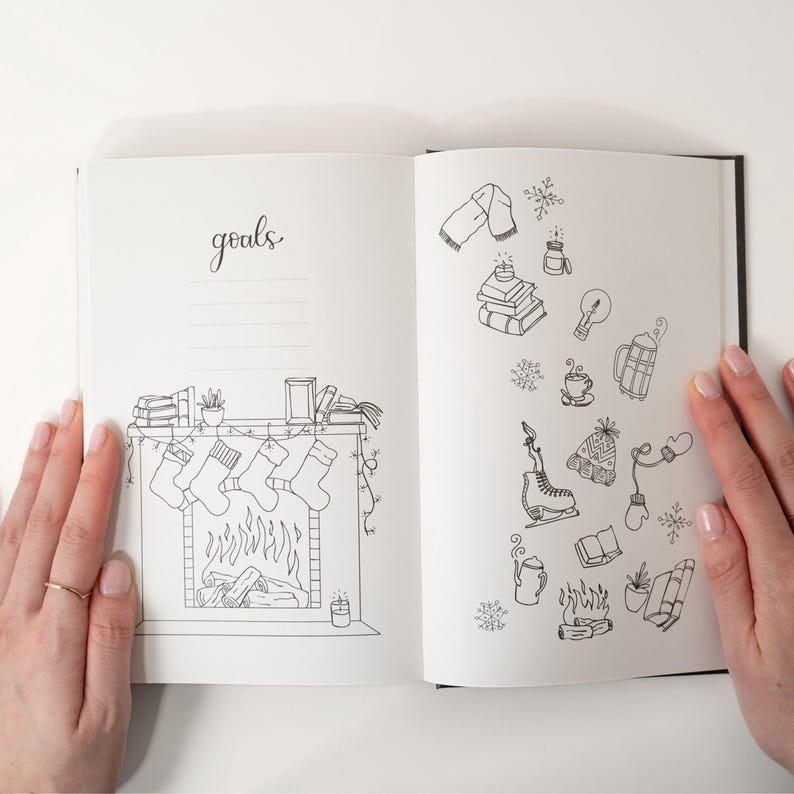 May include: Open journal with black cover, featuring hand-drawn illustrations. One page has the word "goals" and a fireplace with stockings. The other page has winter-themed doodles like a scarf, snowflakes, and ice skates.