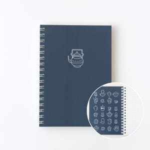 Peut inclure: Un cahier à spirales bleu marine avec une illustration de théière blanche. Le cahier présente une vue rapprochée des pages pointillées et des motifs de théières supplémentaires. Idéal pour la prise de notes et le journal.