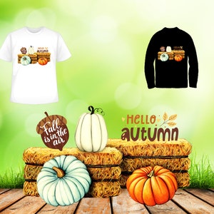 May include: A white t-shirt with a graphic of pumpkins and the text "Hello Autumn". A black long-sleeve shirt with the same graphic. A white pumpkin, a blue pumpkin, and an orange pumpkin are on a stack of hay bales. A sign with the text "Fall is in the air" is next to the white pumpkin. The text "Hello Autumn" is on a hay bale.