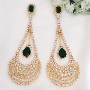 May include: A pair of long gold-tone drop earrings featuring a rectangular emerald-green stone at the top, a central pear-shaped emerald-green stone, and a wide, fan-shaped base encrusted with numerous small, clear, sparkling white crystals.