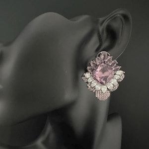 Puede incluir: Primer plano de un pendiente de piedras preciosas rosas y blancas. El pendiente está colocado sobre una cabeza de maniquí negra. El pendiente presenta una gran piedra preciosa rosa cuadrada rodeada de piedras blancas y rosas más pequeñas, creando un diseño floral.