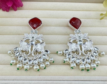 May include: A pair of silver earrings with red gemstone accents. The earrings feature a detailed design of two elephants with intricate floral patterns. The earrings are adorned with white pearls and green beads.