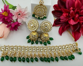 Könnte beinhalten: Ein goldfarbenes Schmuckset mit Halskette, Ohrringen und einem Haarschmuck. Das Set ist mit klaren Steinen, weißen Perlen und grünen Perlen verziert. Die Ohrringe und die Halskette haben ein Design mit hängenden Perlen. Eine rote Blume und rosa Blumen sind im Hintergrund.