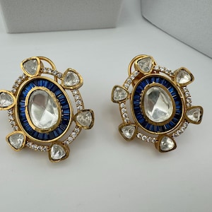 May include: A pair of gold earrings with a large, clear, oval-shaped stone surrounded by a blue and white halo. The earrings are decorated with smaller, clear stones.