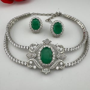 May include: A silver-toned necklace and earring set featuring emerald-green oval gemstones. The necklace has a double-strand design with a central ornate pendant. The earrings are oval-shaped studs. The set is embellished with small, clear gemstones.