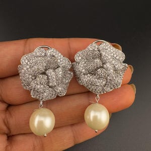 May include: Silver tone flower shaped earrings with a pearl drop. The flower is covered in clear rhinestones.