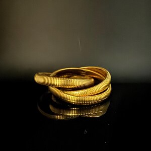 May include: A stack of gold-colored, ribbed bracelets. The bracelets are stacked on top of each other, reflecting on a black surface. The jewelry has a shiny, metallic finish, and the ribbed design adds texture to the bracelets.