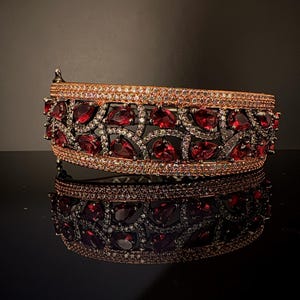 May include: A wide gold-tone bangle bracelet with a delicate, intricate design. The bracelet is adorned with red and clear gemstones, creating a sparkling and elegant look.
