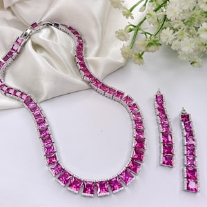May include: A silver necklace and matching earrings set. The jewelry features square, magenta-colored stones set in a silver metal. The necklace and earrings are displayed on a white surface with white flowers in the background.
