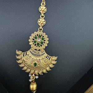May include: Gold-tone maang tikka with green and white stones. The maang tikka has a circular design with a fan-shaped pendant and a pearl and gold tassel.