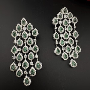 May include: A pair of silver-toned chandelier earrings. Each earring features a cascade of teardrop-shaped green gemstones, outlined with clear crystals. The earrings are displayed against a dark background, highlighting their sparkle and design.