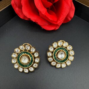 May include: A pair of gold earrings with a circular design. The earrings are adorned with small, clear stones and a larger, central stone. The stones are surrounded by a ring of green stones.