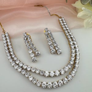 May include: A jewelry set consisting of a double-strand necklace and a pair of matching drop earrings. Each piece features a gold-tone metal setting encrusted with numerous round, clear, faceted stones arranged in a continuous tennis-style design.