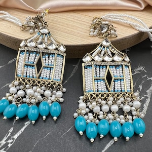 May include: A pair of gold-tone dangle earrings with white and blue beading. The earrings feature a geometric design with a clear crystal embellishment at the top. The earrings are accented with a fringe of white pearls and blue beads.