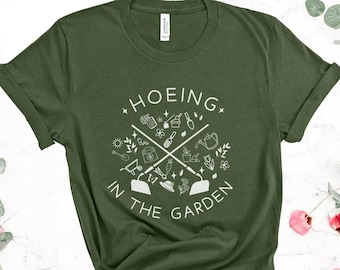Può includere: Una maglietta verde con un design grafico bianco che dice "HOEING IN THE GARDEN" con un cerchio di attrezzi da giardinaggio e piante intorno al testo.