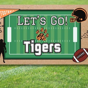 以下が含まれることがあります： 緑色のフィールドデザイン、"LET'S GO!"と"Tigers"の文字、フットボールヘルメット、フットボールが描かれたフットボールをテーマにしたグラフィック。画像には、フットボール選手のシルエットと、"FOOTBALL"と書かれたペナントが含まれています。