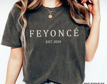 Può includere: Una maglietta grigia scura con il testo "FEYONCÉ EST. 2024" stampato in color oro chiaro. La modella indossa jeans blu e scarpe da ginnastica bianche.