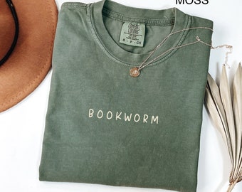 May include: A moss green t-shirt with the text "BOOKWORM" in a light yellow color. The shirt is folded and laid flat on a white surface. A gold necklace with a small pendant is visible on the shirt.
