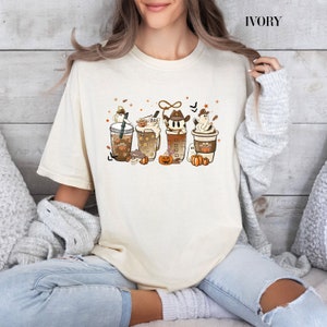 Pode incluir: Uma camiseta cor creme com um gráfico de quatro copos de café temáticos de Halloween. Os copos são decorados com fantasmas, abóboras e a palavra "BOO".
