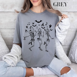May include: A grey t-shirt with a graphic of three skeletons dressed as pumpkins dancing. The skeletons are black and the pumpkins are orange. There are other Halloween-themed graphics around the skeletons, including bats, ghosts, stars, and musical notes.