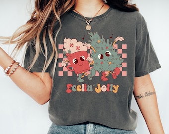 May include: A gray t-shirt with a cartoon illustration of a cup of hot cocoa and a Christmas tree. The text "Feelin' Jolly" is printed in colorful letters below the illustration.