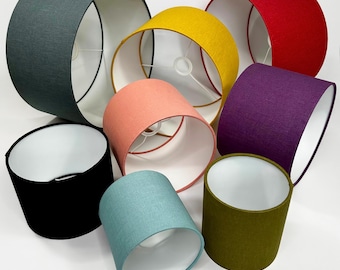 Pode incluir: Uma coleção de sete cúpulas de lâmpada cilíndricas de tecido em várias cores, incluindo cinza escuro, amarelo mostarda, vermelho, roxo, preto, rosa coral e verde oliva. Cada cúpula possui um forro interno branco e uma estrutura de arame.