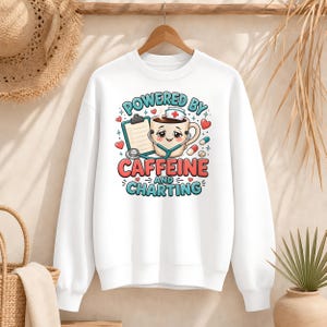 Könnte beinhalten: Ein weißes Langarm-Sweatshirt mit einer Grafik einer Kaffeetasse, die eine Krankenschwesterhaube trägt, dazu ein Stethoskop, ein Klemmbrett und Pillen. Der Text lautet "POWERED BY CAFFEINE AND CHARTING" in blauen und roten Buchstaben.