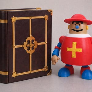 May include: A brown book with gold accents and a decorative figure. The book has a dark brown cover with gold trim and a gold cross design. The figure is red, blue, and tan with a red hat.
