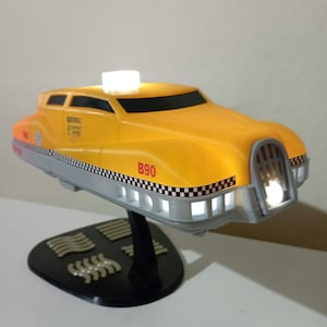 Puede incluir: Un taxi de juguete amarillo con un patrón de tablero de ajedrez blanco y negro, una placa de matrícula roja "B90" y una luz blanca en la parte superior. El taxi está en un soporte negro con una base plateada.