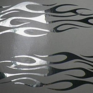 Könnte beinhalten: Zwei Flammenaufkleber, einer über dem anderen, in einem Schwarz-Silber-Farbschema. Die Flammen haben eine stilisierte, längliche Form mit einer reflektierenden Silberoberfläche auf den inneren Teilen. Der Hintergrund ist ein neutrales Grau.