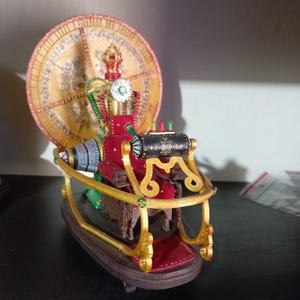 Op de afbeelding: Gedetailleerd steampunk-beeldje met een rode zitting, gouden accenten en een grote, cirkelvormige achtergrond. Het beeldje heeft een zwart cilindrisch onderdeel en ingewikkelde details, met een slee-achtige basis.